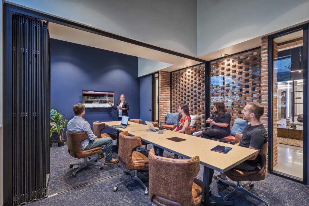 A modern huddle room with a team of five professionals engaged in a presentation. The room features brick accent walls, a large table with laptops, comfortable swivel chairs, and a presentation screen. The ambiance is professional yet inviting, fostering collaboration and communication