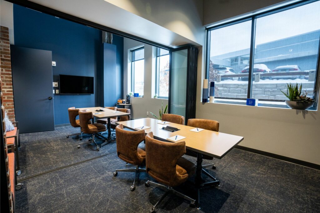 Spacious huddle room with a modern design, featuring a large window with a city view, a flat-screen tv for presentations, and multiple tables with plush swivel chairs. The room is ready for collaborative meetings, with a balance of comfort and technology.