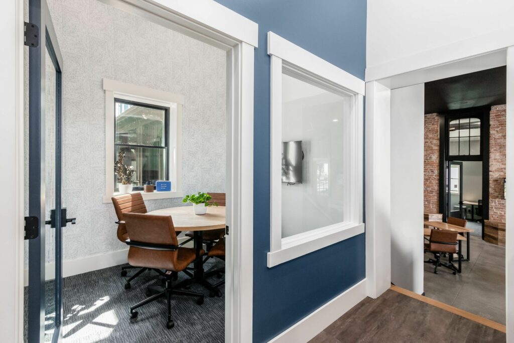 View into a small, stylish huddle room through an open door, revealing a round table with leather chairs and modern decor. The room is well-lit with natural light from a window, creating a warm and inviting atmosphere for small team meetings or private work.