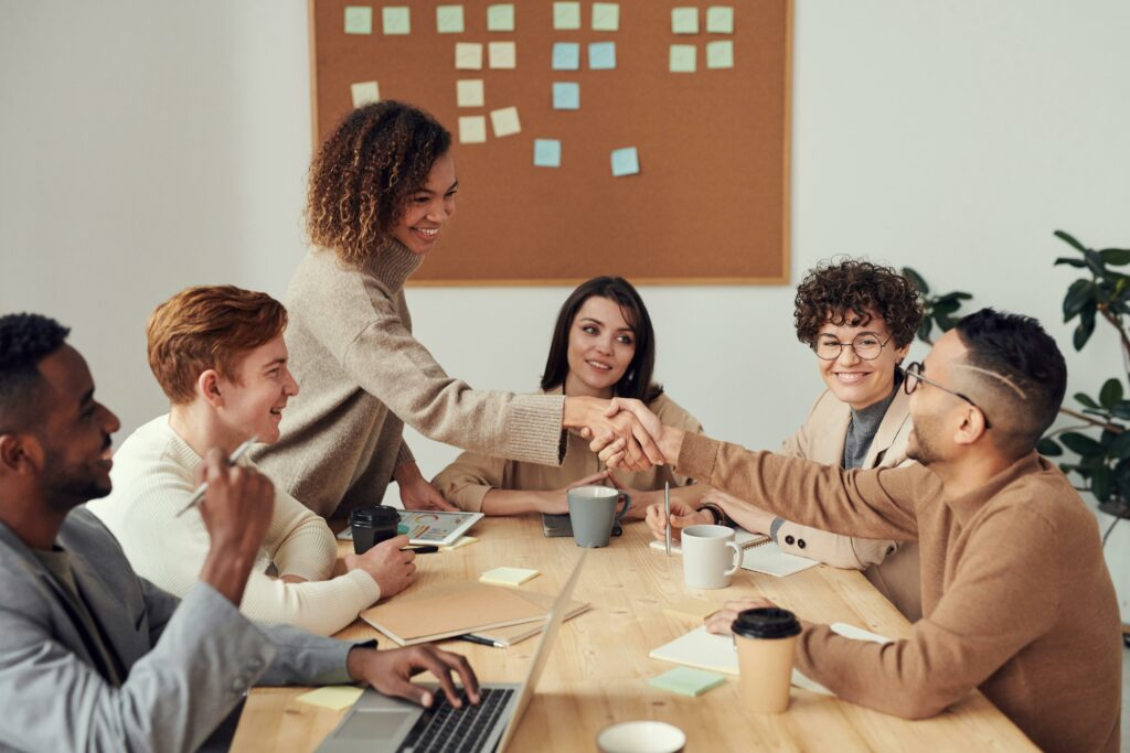 A team of diverse professionals engaging in a collaborative handshake over a wooden table in a huddle room, with laptops, coffee mugs, and sticky notes around them. They display a friendly atmosphere with smiling faces, embodying teamwork and successful agreement.