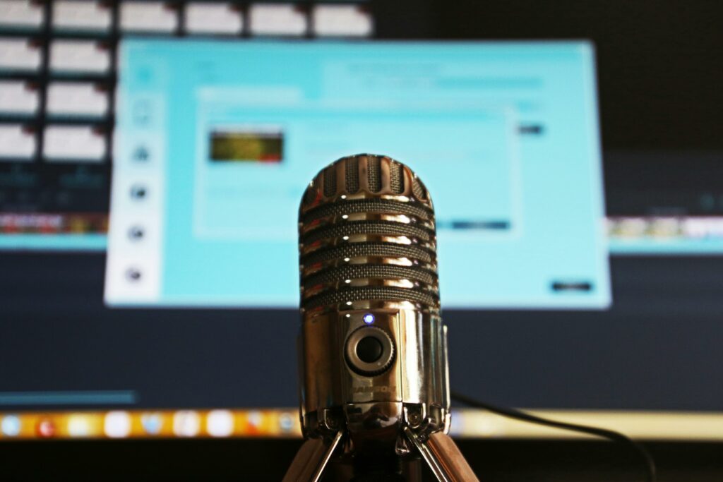 A classic microphone in the foreground with a focus on the audio equipment, suggesting a huddle room setup for a podcast recording or audio conferencing, with audio production software visible on the computer screen in the soft-focused background.